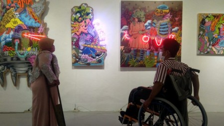 Foto pengguna kursi roda dan pengguna tongkat sedang melihat karya lukis yang tergantung didinding